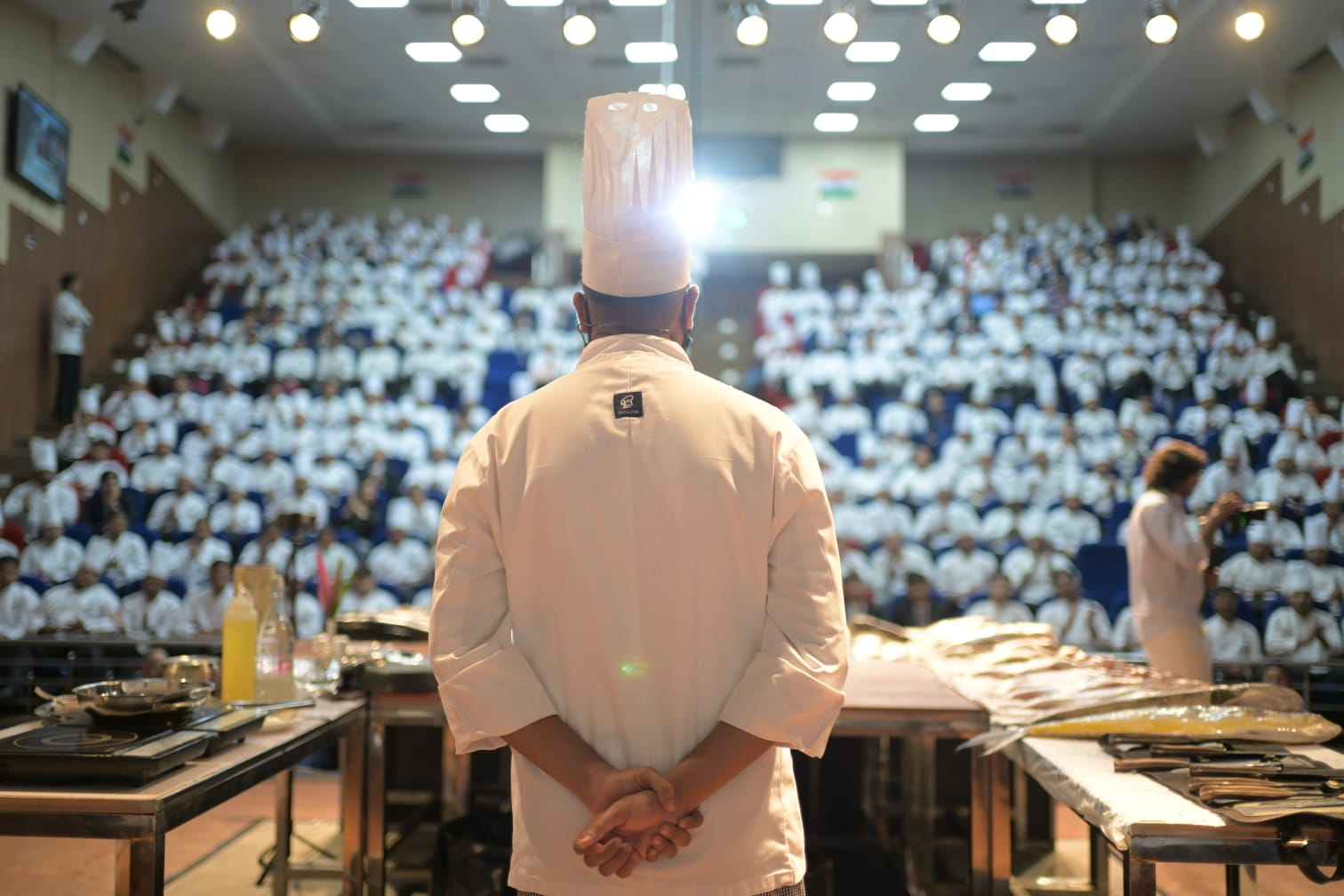 Chef Mohammad Husain standing with his back to the camera, facing a large student audience, with fresh fish laid out on tables at the IHM Bhopal seafood workshop